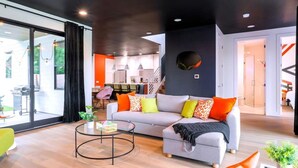 A modern living room with a grey sofa, colorful cushions, a glass coffee table, and a large window. - Group Getaway With Chess Garden and Firepit (Austin)