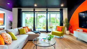 A modern living room with a sofa, armchair, and coffee table, large windows with curtains, and a TV on the wall. - Group Getaway With Chess Garden and Firepit (Austin)
