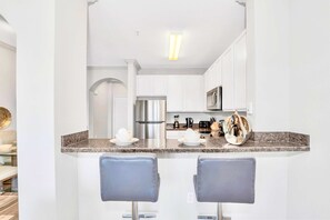 Uma cozinha moderna com armários brancos, eletrodomésticos de aço inoxidável e bancada de granito. Há dois banquetes sob a bancada.