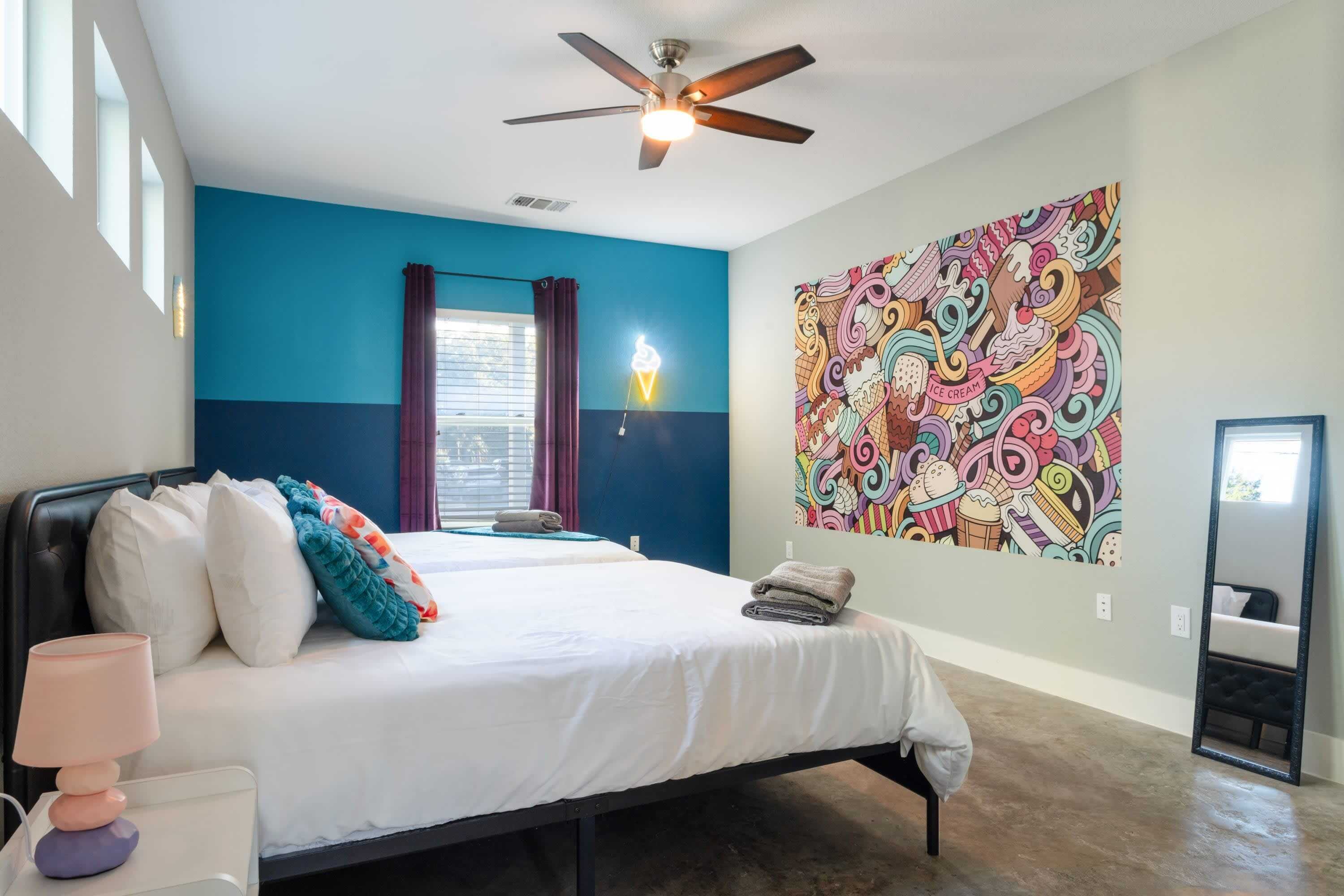 A bedroom with a bed, pillows, a bedside lamp, a ceiling fan, a window with curtains, and a colorful wall mural.