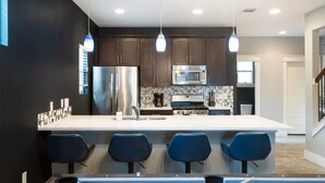 A modern kitchen with dark cabinets, stainless steel appliances, a white island with bar stools, and pendant lighting. - Upstay Entertainment Hub w Games & Outdoor Firepit (Austin)
