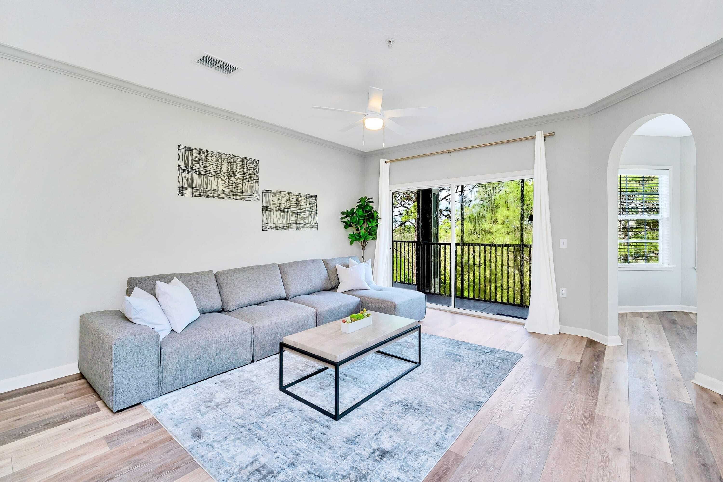 A modern living room with a grey sectional sofa, a wooden coffee table, and a large window with a view of greenery.