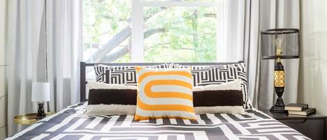 Ein Bett mit einer schwarz-weiß gestreiften Bettdecke, einem einzelnen orange-weißen Kissen und einer schwarzen Lampe auf einem Nachttisch.