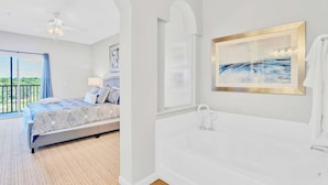 A bedroom with a bed, a large window, a ceiling fan, and a bathroom with a bathtub and a framed picture on the wall. - Upstay: Condo Near Disney w Games, Pool (Winter Garden)