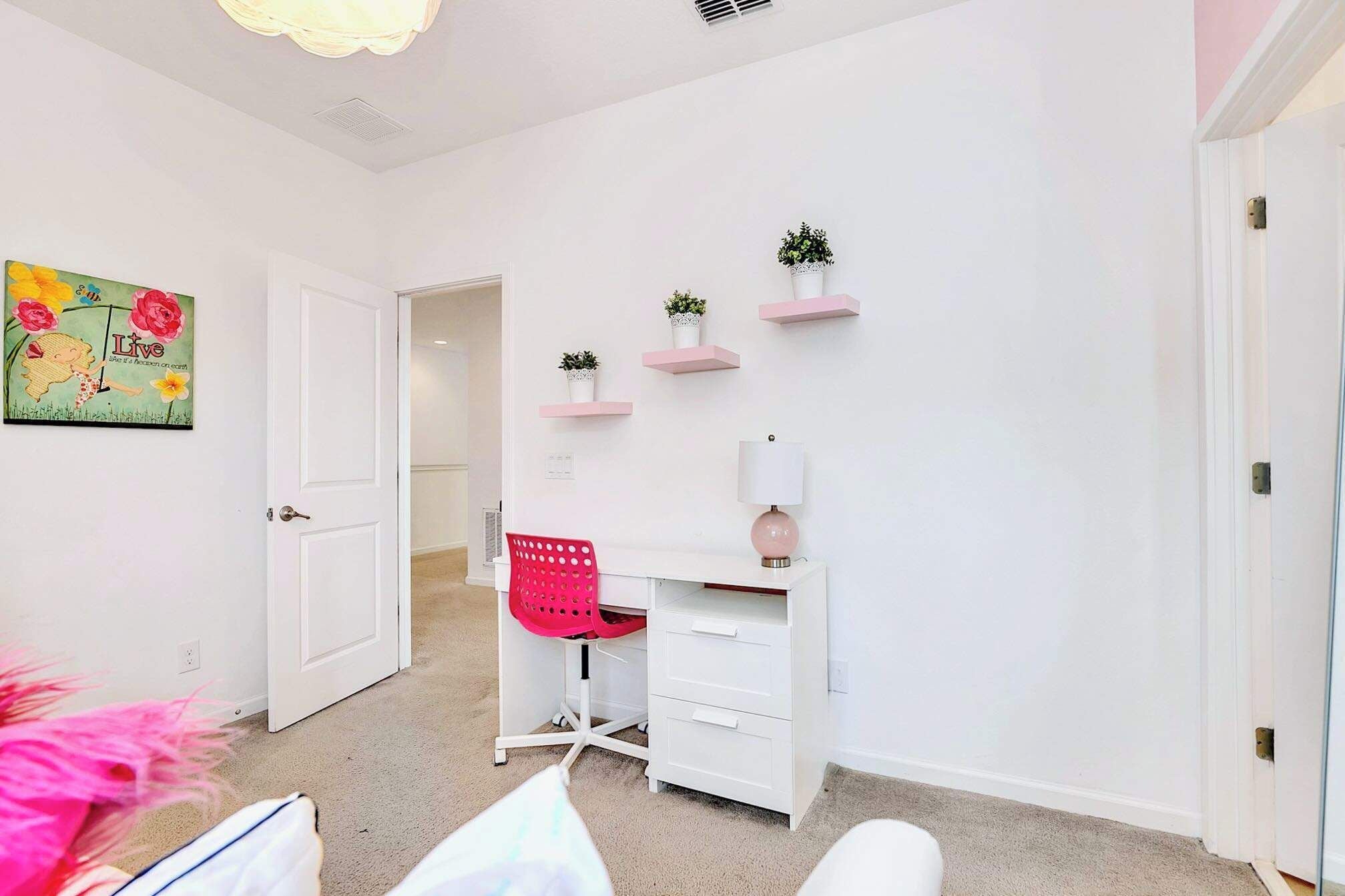 Ein Zimmer mit einem weißen Schreibtisch, einem roten Stuhl und einer weißen Tür. Rosa Regale mit Pflanzen befinden sich an der Wand.