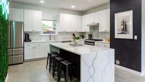 A modern kitchen with white cabinets, a stainless steel refrigerator, a large island with stools, and a black accent wall. - East Austin Pool and Hot Tub Retreat Near Rainey (Austin)