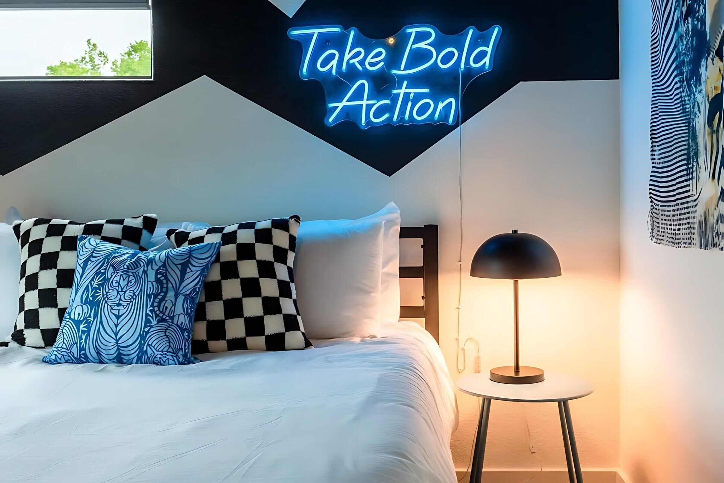 A bedroom with a bed, checkered pillows, a neon sign, a lamp, and a wall with a cityscape mural.