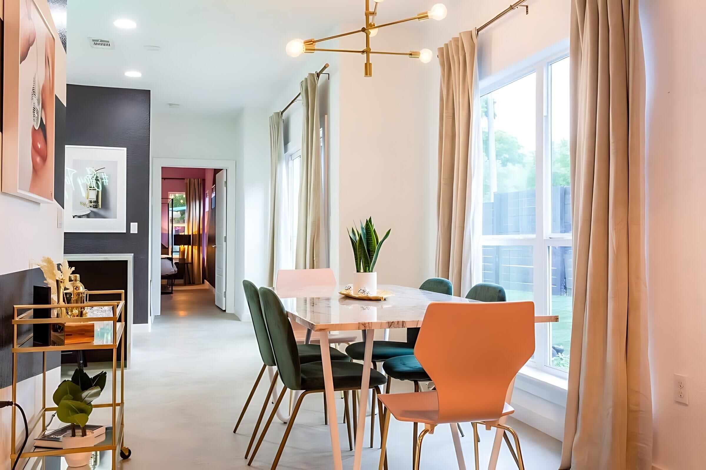 A modern dining area with a marble table, green and orange chairs, a potted plant, and a view of the outdoors.