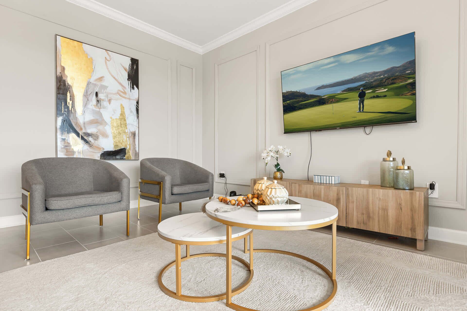 Una sala de estar moderna con dos sillones grises, una mesa redonda para café, un cuadro abstracto grande, un televisor mostrando un campo de golf y un mueble de madera con adornos.