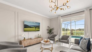 A modern living room with a large TV, comfortable seating, a coffee table, and a view of a pool through large windows. - Upstay: Poolside Mansion w/ Themed Arcade & Cinema (Four Corners)