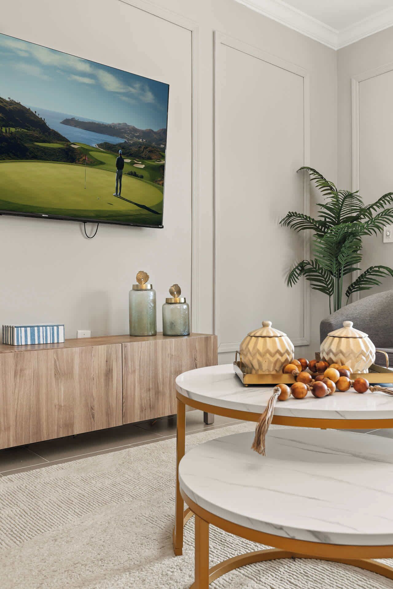 Un salon moderne avec un téléviseur à écran plat fixé au mur, une console en bois ornée de vases décoratifs, une table ronde en marbre sur laquelle repose un bol de noix, et une grande plante verte.