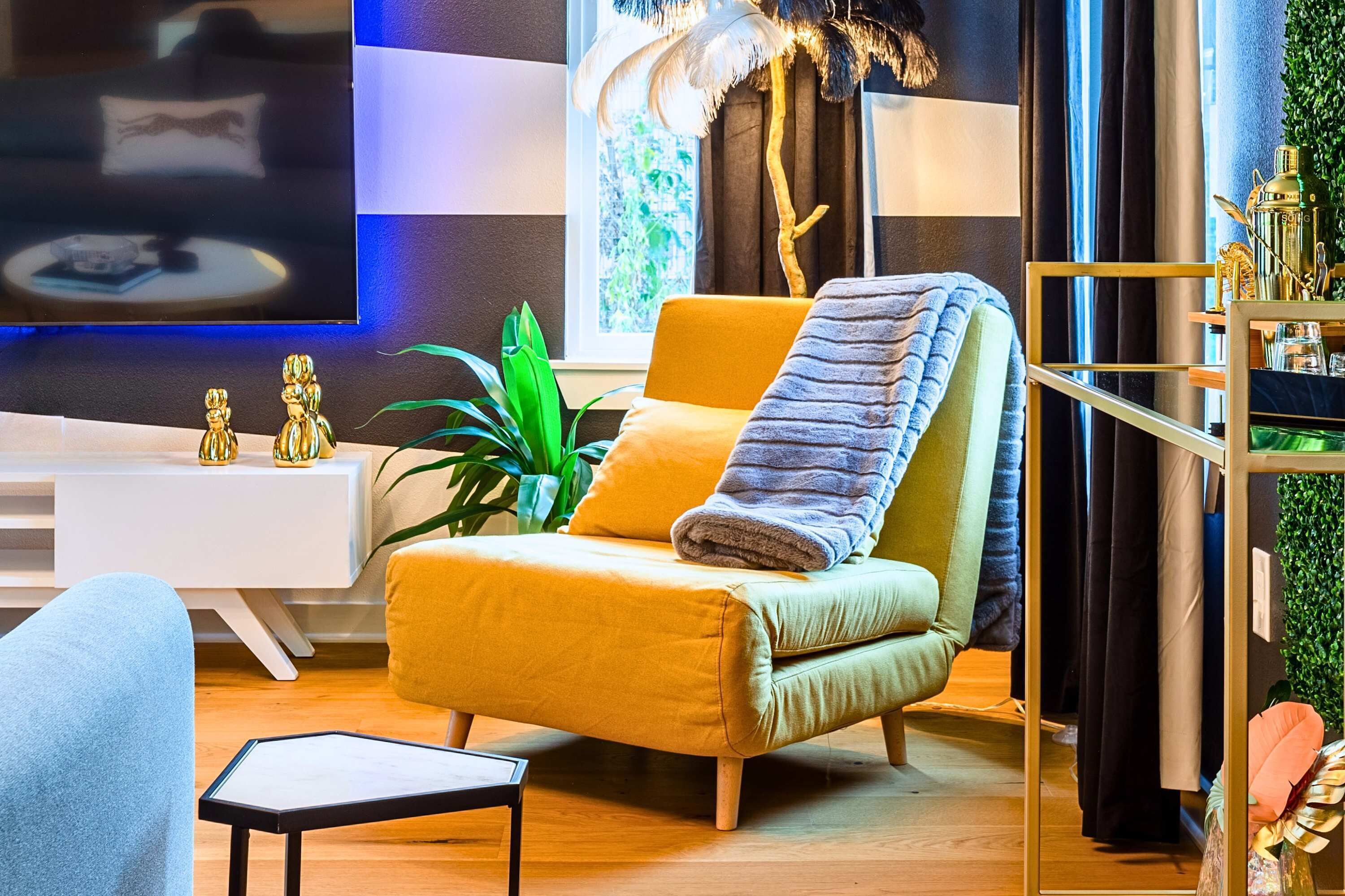 A modern living room with a yellow armchair, a white side table, a flat-screen TV, and a large window with curtains.