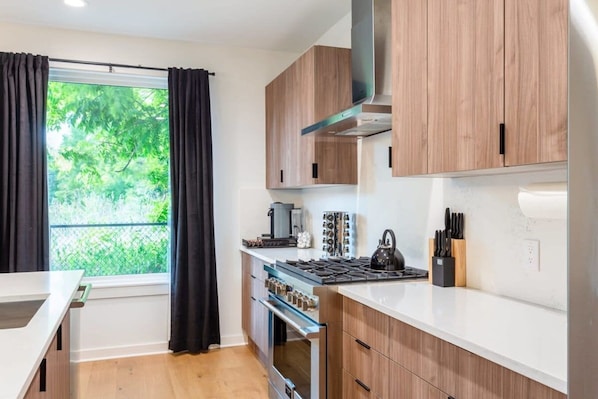 A modern kitchen with wooden cabinets, a gas stove, and a window with curtains. - Stylish Clubhouse Retreat w/ Hot Tub Near Downtown (Austin)