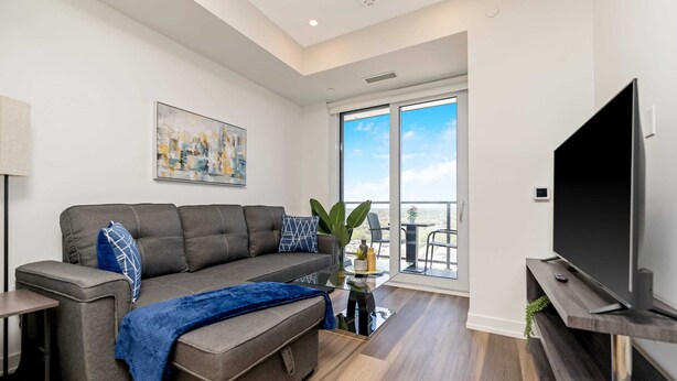 A modern living room with a grey sectional sofa, a flat-screen TV on a wooden stand, and a large window offering a view of the outdoors. - Stylish 1BR Condo w Balcony & Skyline Views (Kitchener)