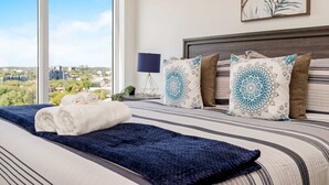 A neatly made bed with a dark blue blanket and rolled white towels, a bedside lamp, patterned pillows, and a city view through large windows. - Stylish 1BR Condo w Balcony & Skyline Views (Kitchener)