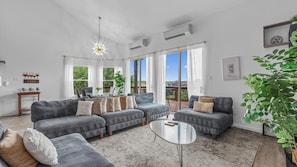A spacious living room with a large sectional sofa, a coffee table, and a dining area with a glass table and chairs. There are large windows providing natural light and a view of the outdoors. - Large 6BR Lakefront Home With Private Pool & Views (Lakeway)