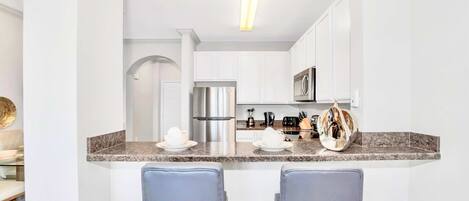 Una cocina moderna con armarios blancos, electrodomésticos de acero inoxidable y encimera de granito. Hay dos taburetes de bar debajo de la encimera.