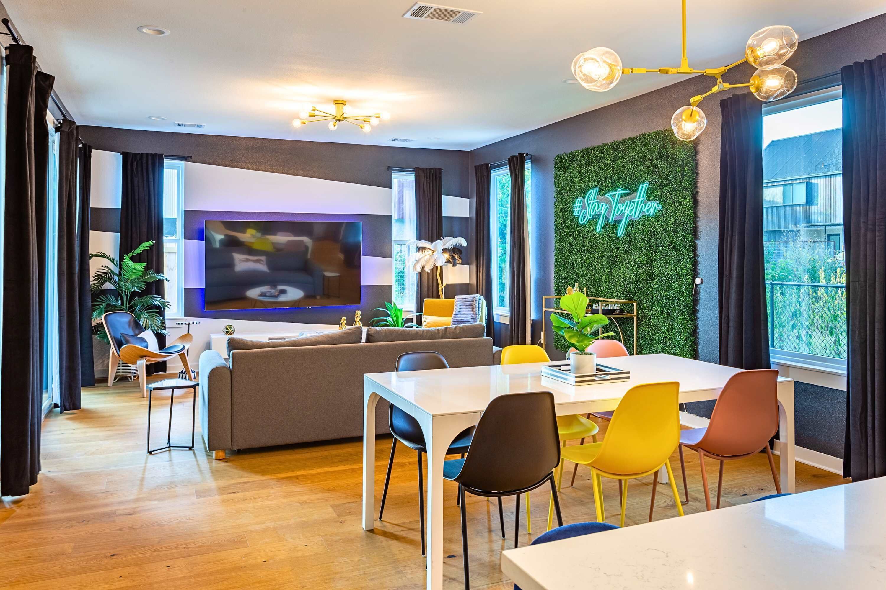 Een moderne woonkamer met een flatscreen-tv, een eethoek met kleurrijke stoelen, een bank en een groene muur met een #Together bord.