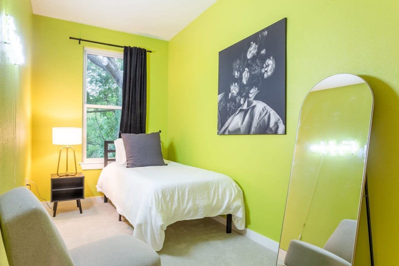 A room with a bed, a bedside table with a lamp, a chair, a large mirror, and a window with curtains.