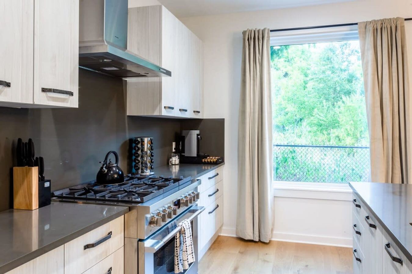 A modern kitchen with a gas stove, oven, and a coffee machine. There is a window with curtains and a view of greenery outside.