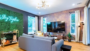 A modern living room with a grey sofa, a flat-screen TV, a green wall with a 'Stay Together' sign, and large windows.