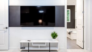 A modern living room with a flat-screen TV mounted on the wall, a white console table with a plant, and a door leading to a bathroom.