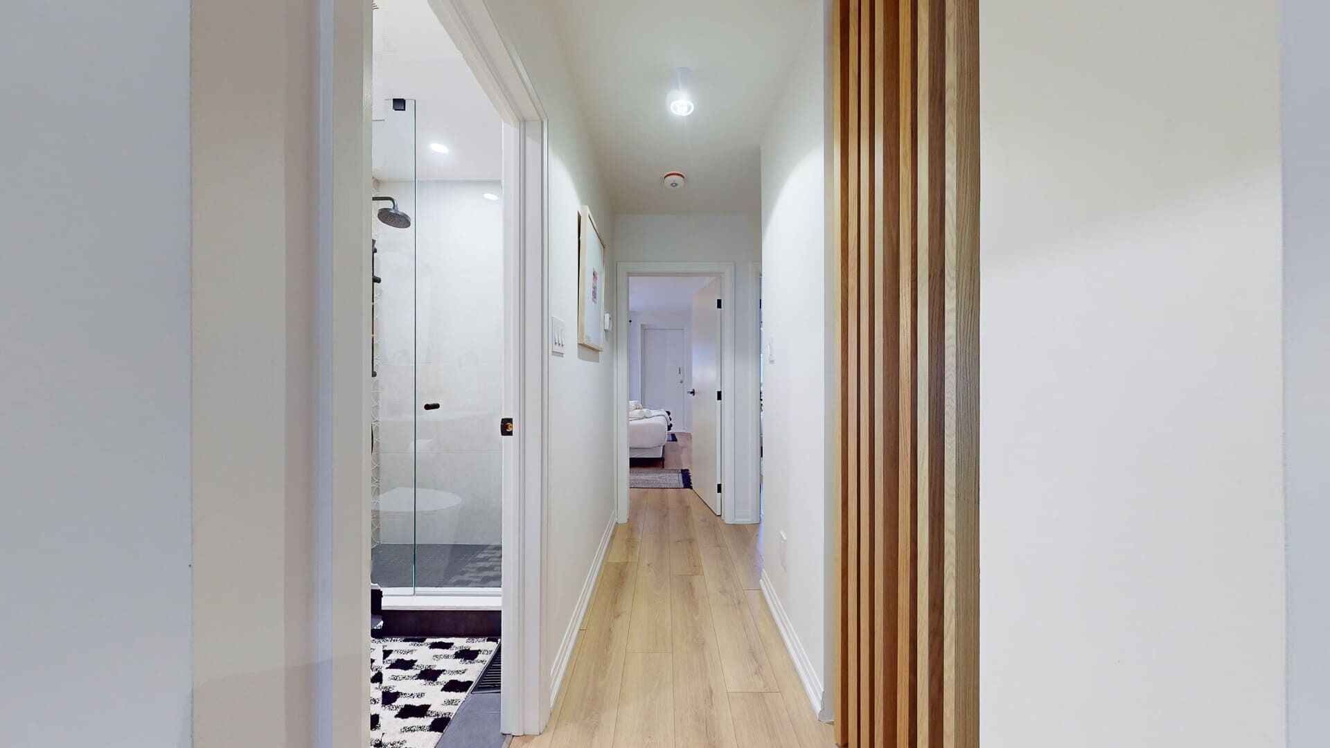 A narrow hallway with a glass shower enclosure, a bed in the background, and a wooden panel on the right wall.