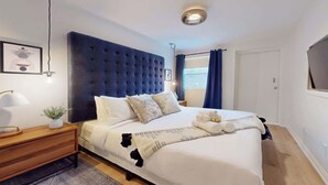 A modern bedroom with a tufted headboard, a neatly made bed with white linens, a bedside table with a lamp and a small plant, and a window with blue curtains. - Casa Hotels Elegant 2BR Suite in Leslieville (Toronto)