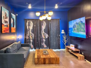 A modern living room with a blue accent wall, a large flat-screen TV, a wooden coffee table, a grey sofa, and a unique black curtain with a white abstract design. - Ultimate 10br Group Getaway6min to Rainey Street (Austin)