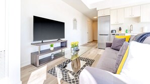 A modern living room with a flat-screen TV, a glass coffee table, and a sofa with cushions. - High Rise 1BR Building With Gorgeous Views (Kitchener)