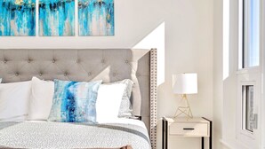 A neatly arranged bedroom with a large bed, a bedside table with a lamp, and a window with curtains. - High Rise 1BR Building With Gorgeous Views (Kitchener)