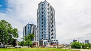 A modern high-rise building with a glass facade, surrounded by a well-maintained lawn and a few trees. - High Rise 1BR Building With Gorgeous Views (Kitchener)
