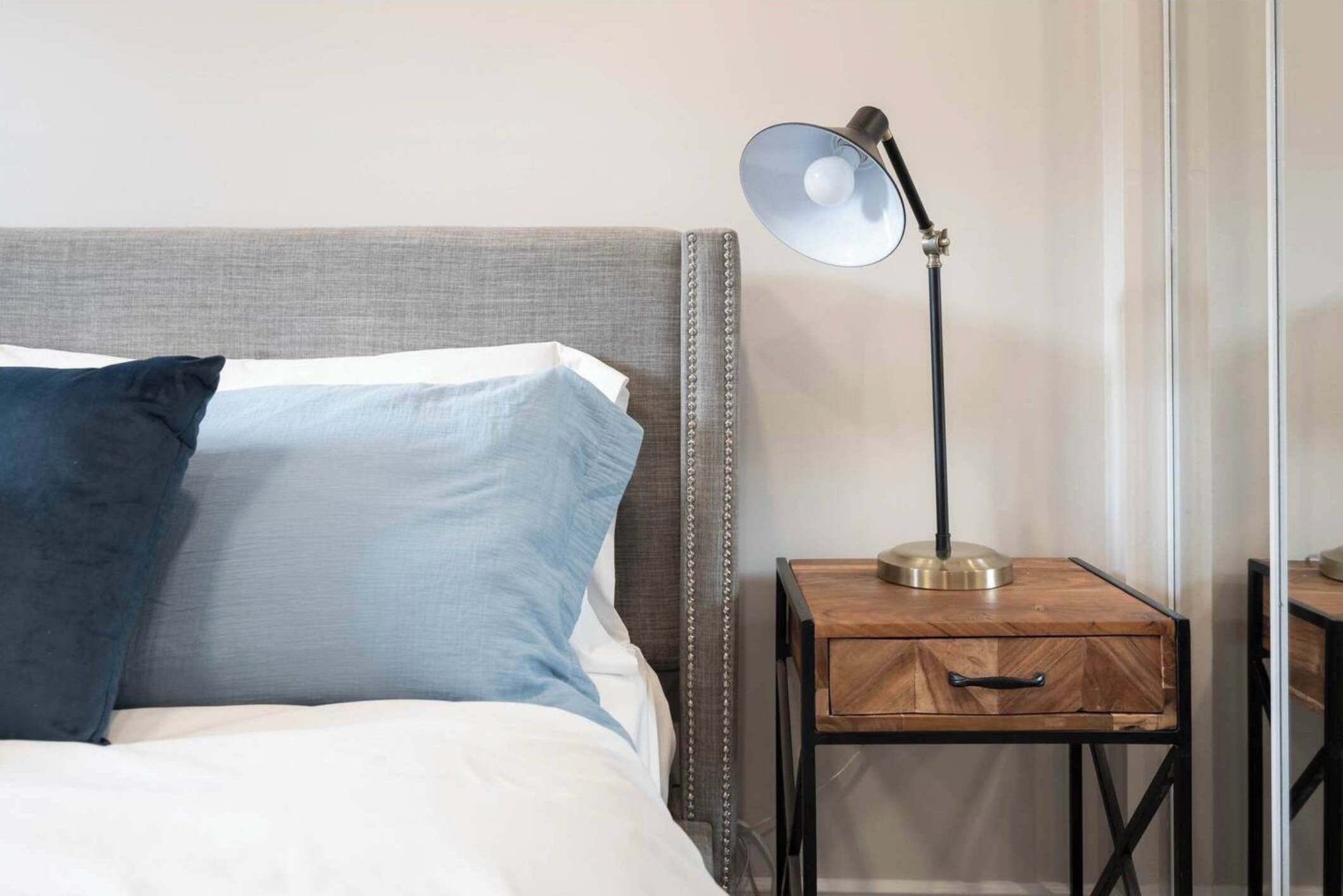 A neatly made bed with a grey headboard, a blue pillow, and a white pillow. There is a small wooden nightstand with a brass lamp beside the bed.