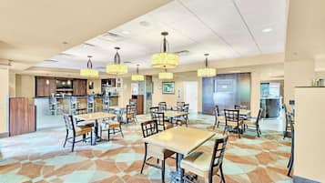 Uma sala de jantar espaçosa com várias mesas e cadeiras, cozinha ao fundo e iluminação pendente.