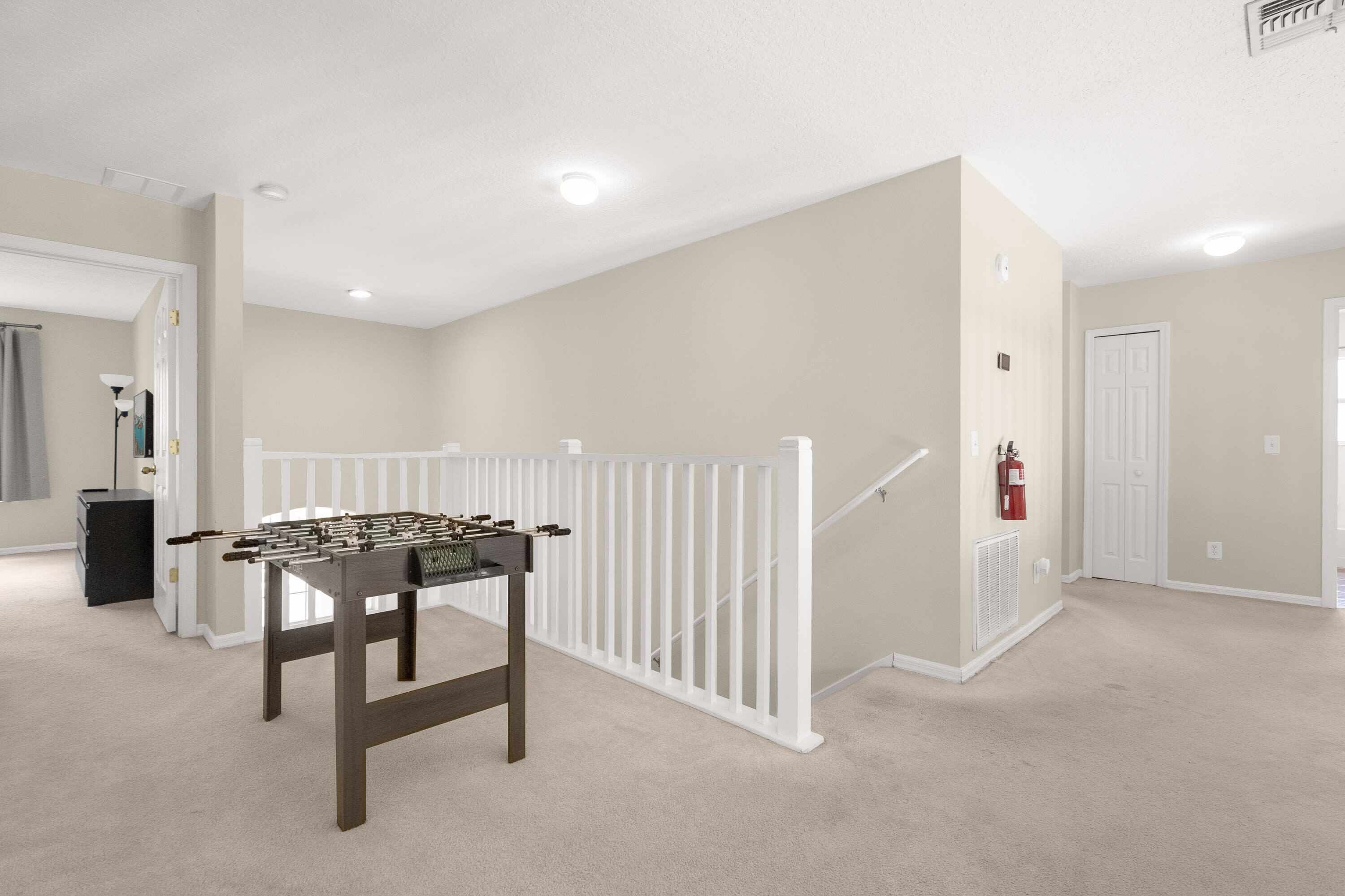 A room with a foosball table, a white railing, a fire extinguisher, and a staircase.
