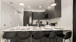 A modern kitchen with a central island, dark cabinets, and a staircase leading to another level. - Contemporary 4BR Townhome With Pool & Themed Rooms (Clermont)