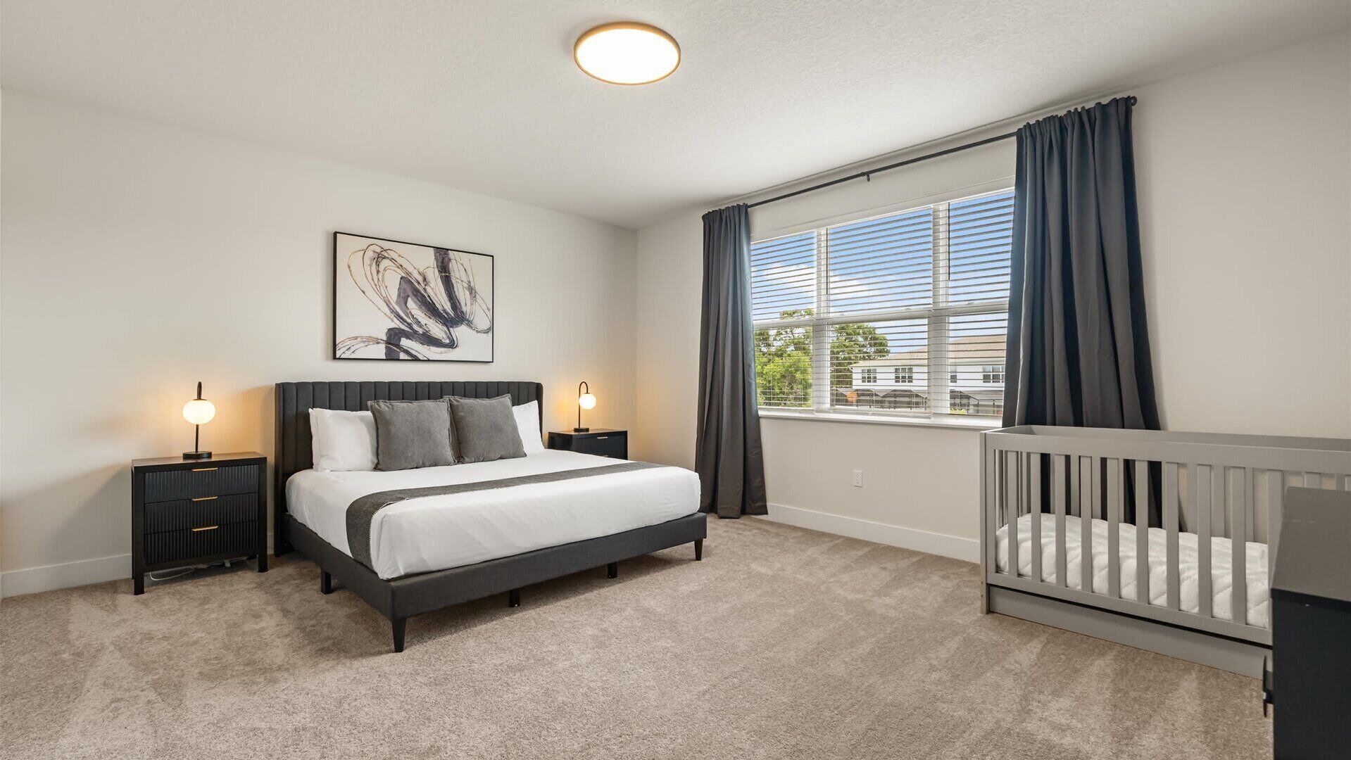 A bedroom with a bed, bedside tables, a crib, a window with curtains, and a ceiling light.