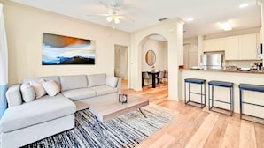 A modern living room with a sectional sofa, a coffee table, and a painting on the wall. There is a kitchen area with a refrigerator, a microwave, and a dining table with chairs. - Upstay 3BR Suite w Pool, Sauna & Gamesnear Disney (Winter Garden)