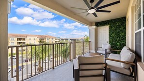 Un balcon doté d’un ventilateur de plafond, un coin salon avec deux fauteuils et un canapé, et une vue sur un quartier résidentiel avec des immeubles et des voitures garées.