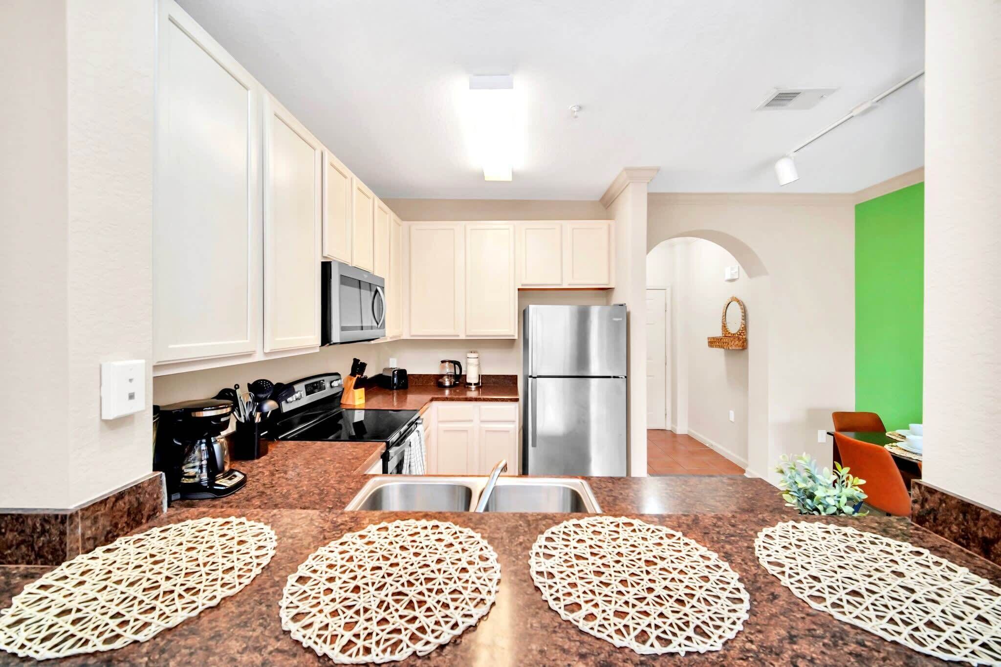 A modern kitchen with white cabinets, a stainless steel refrigerator, and a microwave. There are two sinks and a coffee maker on the counter.