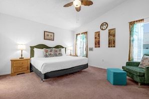 Une chambre à coucher avec un lit, des tables de chevet, un fauteuil vert et une fenêtre avec des rideaux.