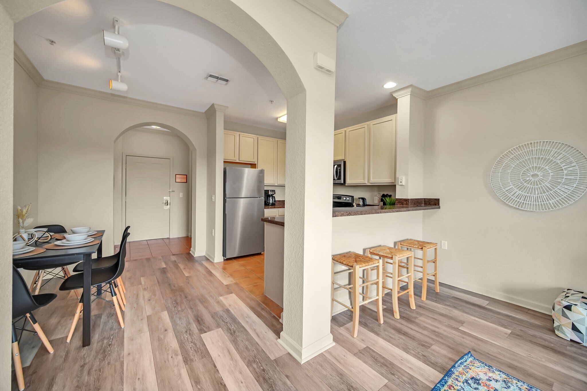 Una cocina moderna con refrigerador integrado, microondas y cafetera. Hay una zona de comedor con mesa y sillas, además de un pequeño bar con taburetes.