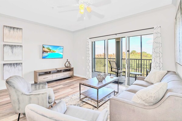A modern living room with a sofa, armchairs, a coffee table, and a TV. There is a balcony with a view of trees and a railing.