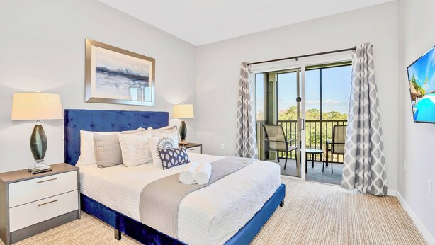 A hotel room with a large bed, two bedside tables, a painting, and a view of the outdoors through a sliding glass door. - Upstay - Large Condo Near Disney w Pool and Gym (Winter Garden)