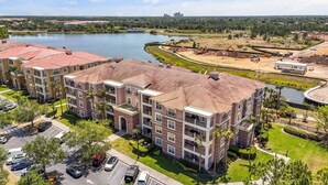 Un edificio residencial de varios pisos con techo de tejas rojas, rodeado por un estacionamiento y una masa de agua.