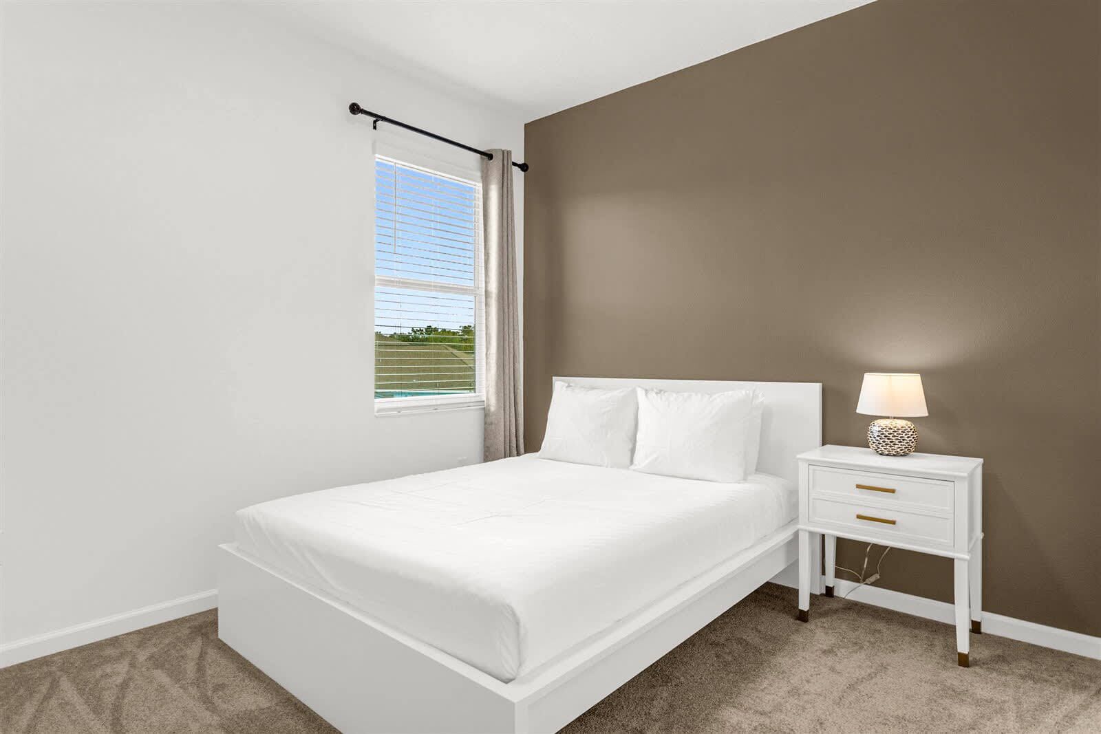 Un dormitorio con una cama blanca, una mesita de noche con lámpara y una ventana con persianas.