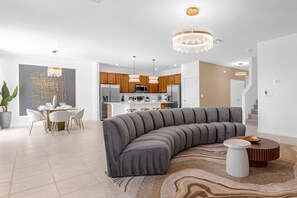 A modern living room with a curved grey sofa, a dining area with white chairs, and a kitchen with wooden cabinets and stainless steel appliances. - Upstay - Mansion w/ Kids Theme Rooms, Pool, Cinema (Kissimmee)