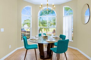 Un coin repas avec une table ronde, quatre chaises et un lustre. Il y a trois grandes fenêtres à cadres blancs, donnant sur un paysage verdoyant.