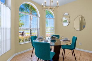 Un coin repas avec une table ronde, des chaises de couleur turquoise et une vue sur les palmiers à travers de grandes fenêtres.