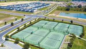 Un gran complejo deportivo con varias canchas de tenis, un área de estacionamiento y un barrio residencial al fondo.